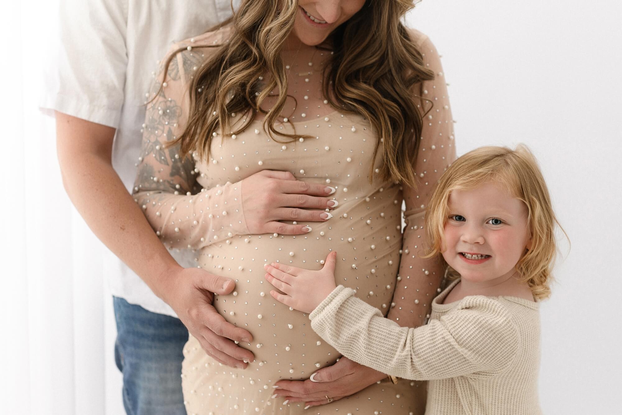 Pregnant mother in pearl-embellished dress with partner’s hands on belly and smiling toddler hugging her during maternity photos with partner and siblings in Portland, Oregon.