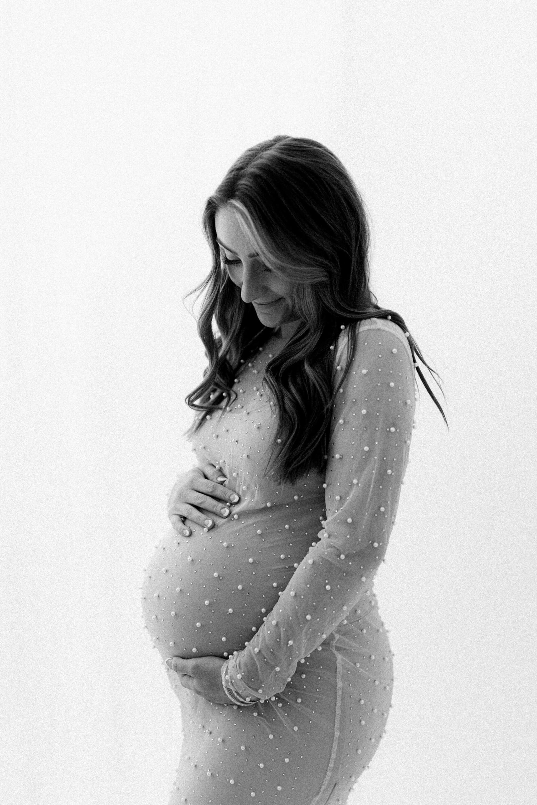 Black-and-white portrait of pregnant woman in sheer pearl dress cradling her belly during maternity photos with partner and siblings in Portland, Oregon.